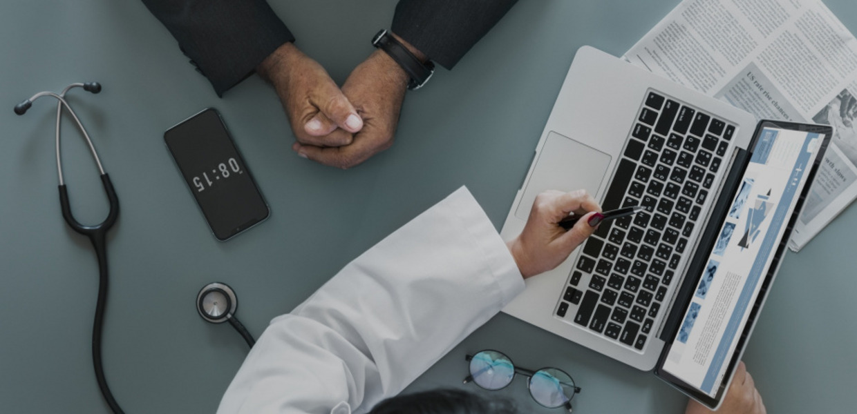 (© rawpixel/pixabay.com - CC0, Public Domain, https://creativecommons.org/publicdomain/zero/1.0/deed.de) Arm im weißen Kittel mit Händen auf dem Notebook und die verschränkten Arme einer anderen Person von oben an einem Tisch sitzend. Dort liegt auch ein Stethoskop